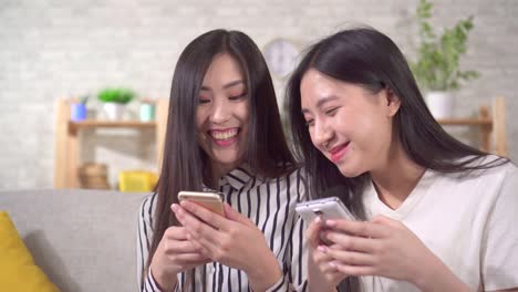 two happy young asian girls with smartphones are sitting on the sofa in the living room close up