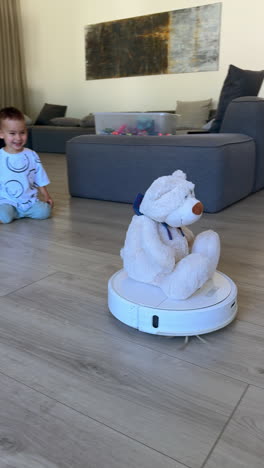 cute toddler boy looks with interest on the big white teddy sitting on the spinning robot vacuum cleaner. kid laughs cheerfully watching device work. vertical video.