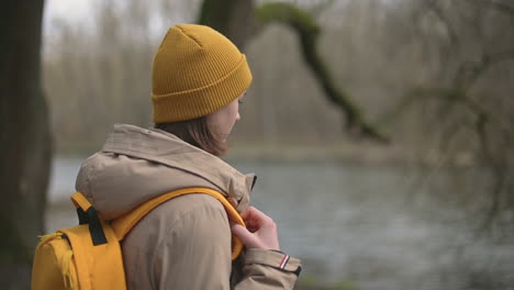 a young girl in a forest that wears a yellow wool cap and a yellow backpack walks towards the lake