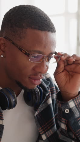 man with glasses works on laptop in library. young black guy with headphones adjusts spectacles sitting at computer in public hall. student technology