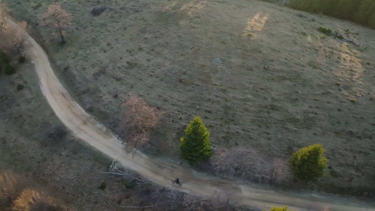 un camino de tierra sinuoso con un ciclista a través de un bosque y prado durante la hora dorada, vista aérea