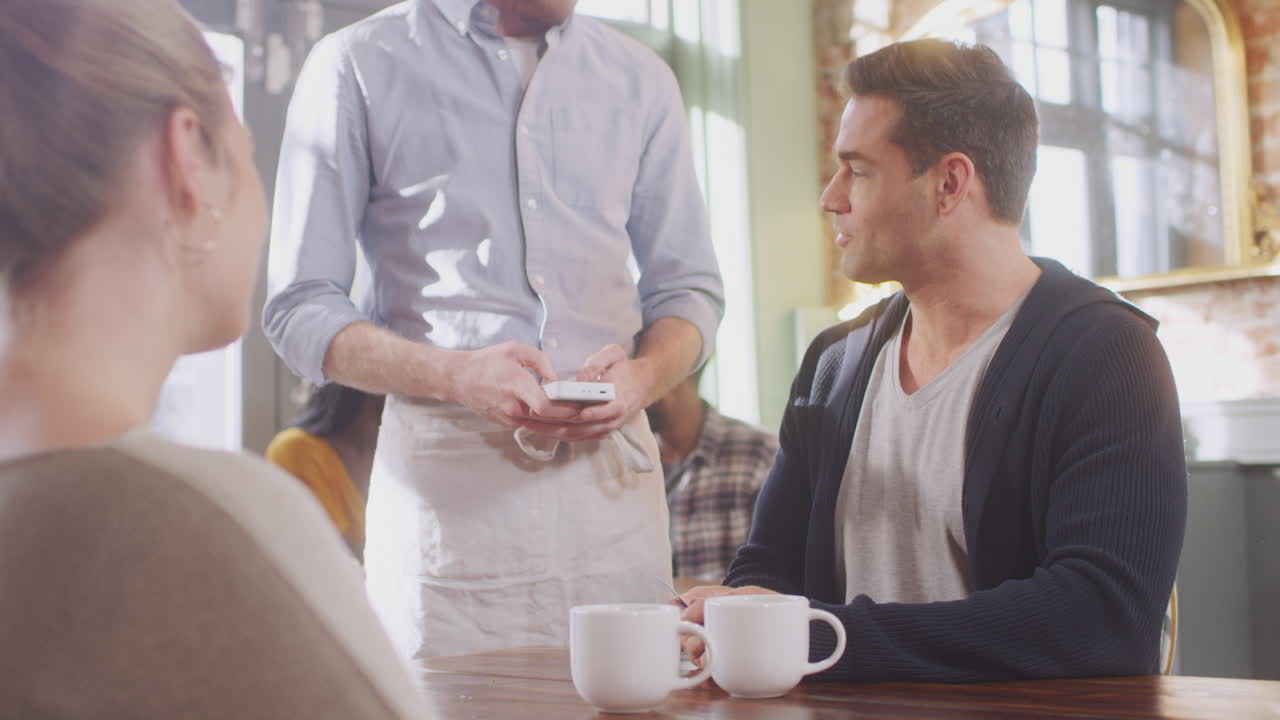 Couple In Coffee Shop Making Contactless Payment With Card For Drinks To Male Waiter