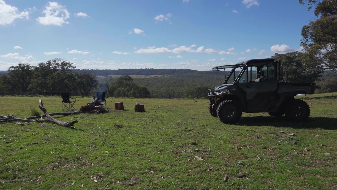 Bred rörlig gimbalbild av en avlägsen lägerplats med lägereld och ett robust jordbruksfordon med utsikt över böljande busklandskap under en klarblå himmel på landsbygden i New South Wales, Australien