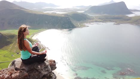 a young, white female hiker is sitting down and meditating on a rock on the edge of a cliff on top of mannen mountain, overlooking haukland beach during a warm and sunny summer day in lofoten, norway