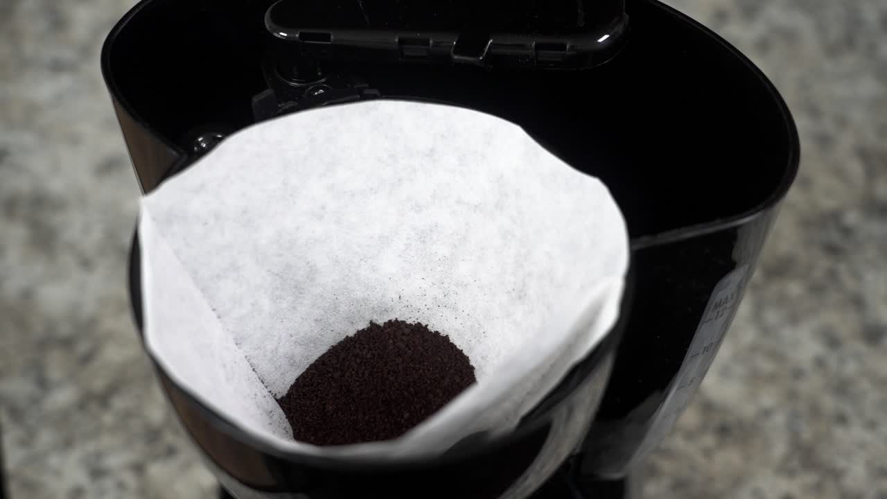Morning routine showing a person pouring fresh ground coffee into a paper filter to prepare a pot of hot drip coffee for breakfast