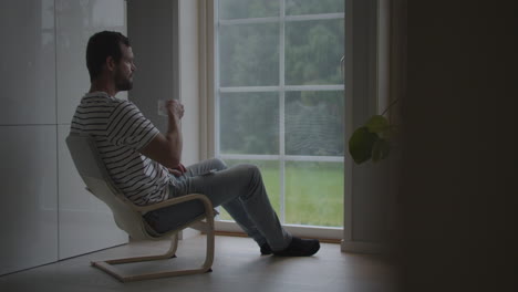 large nordic man sits by window drinking coffee looking at rain outside