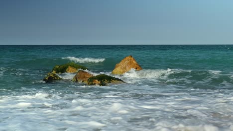 small waves crash over some rocks just of a sandy mediterranean beach