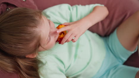 overhead view of young girl relaxing on couch wearing mint clothes taking big bite from juicy fruit, holding it with both hands, cozy indoor atmosphere, casual mood, natural lighting