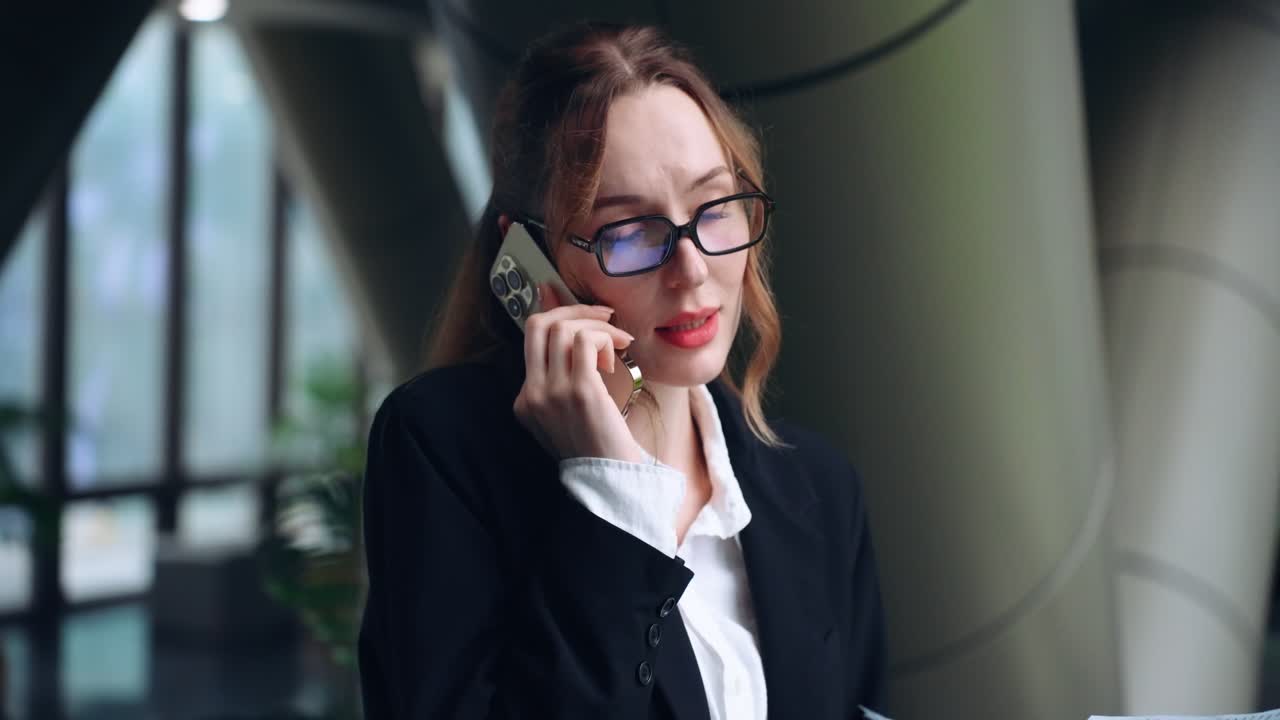 Focused corporate executive in suit and glasses holds paper work and talks on cell phone in modern office environment with concerned expression