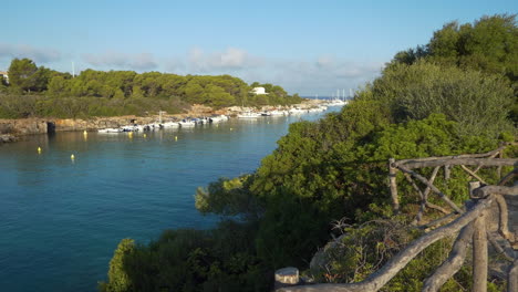 Wide-view-from-a-high-point-behind-the-wooden-barriers-of-the-Cala-Santandria-in-Menorca-just-after-dawn,-blue-seawater,-surrounding-rocks,-boats,-green-trees