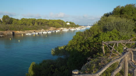 wide view from a high point behind the wooden barriers of the cala santandria in menorca just after dawn, blue seawater, surrounding rocks, boats, green trees