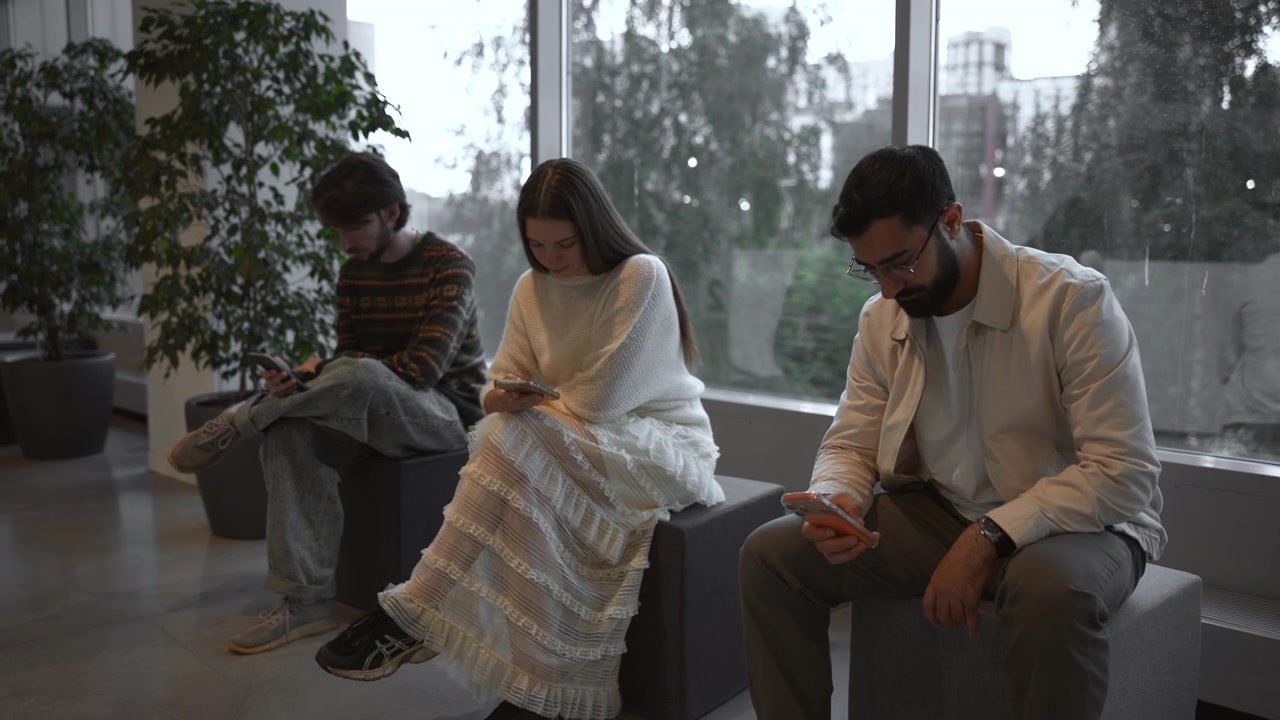 Tre voksne sitter på kubebenker i en moderne lobby, bøyd fremover med øynene på håndholdte smarttelefoner, mens regnflekkete glass, potteplanter og mykt dagslys former en rolig urban ventescene