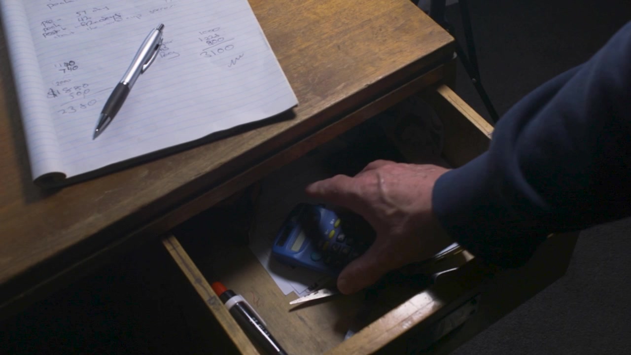 Adult hand opens a wooden desk drawer under handwritten notes and pulls out a small blue calculator.