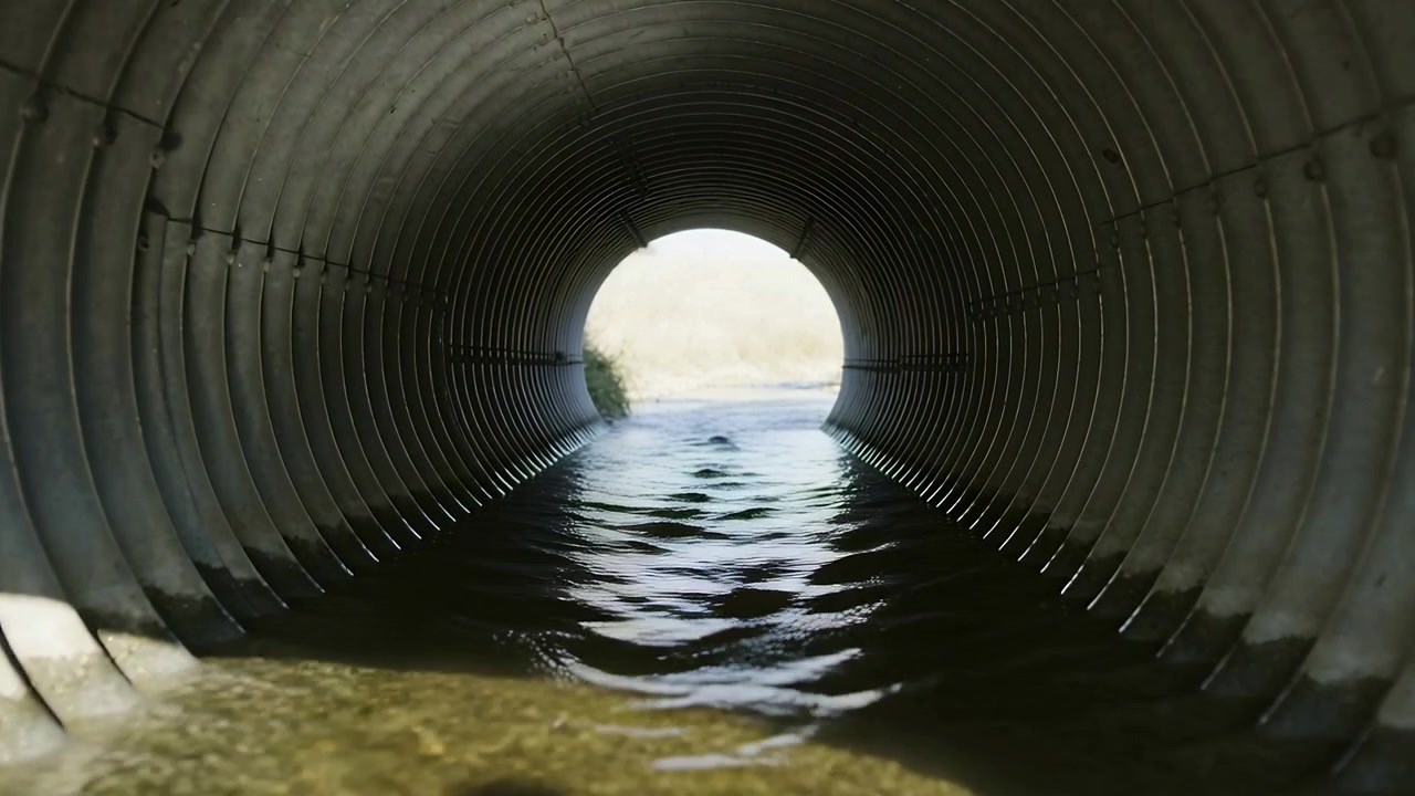 Agua fluyendo entrando desde un campo en una tubería corrugada, drenando hacia la cámara, ondeando con la luz.