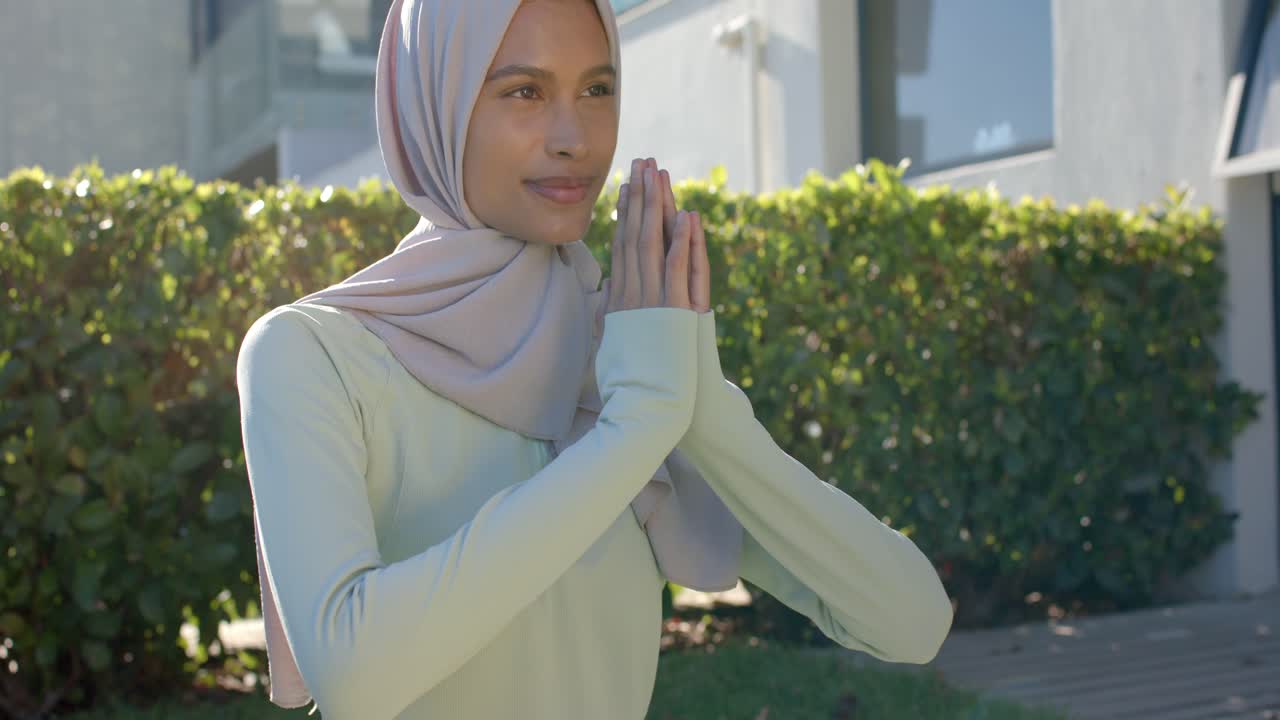 Vídeo vertical: Mulher de hijab claro e blusa clara, com as mãos em oração, ajustando-se em um cumprimento em um gramado.