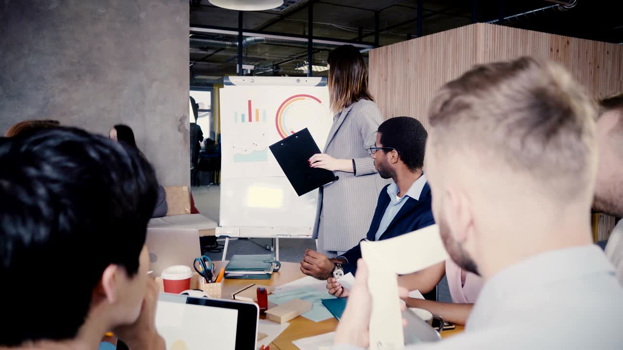 여성 ceo가 팀에 비즈니스 프로젝트를 설명하고, 플립 차트에서 데이터를 설명합니다. 다민족 사무실 직원 이사회 회의 4k