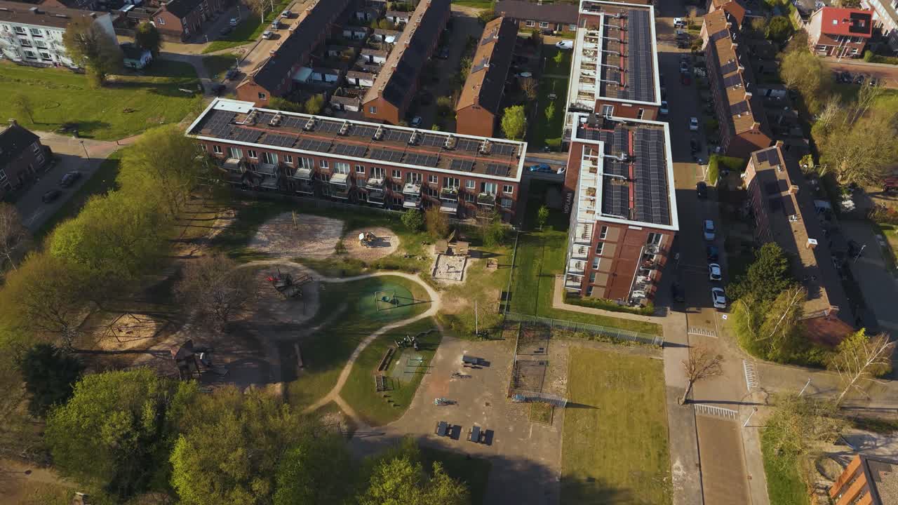 Drone footage of a peaceful suburban neighborhood in Leident, Netherlands, showing brick housing blocks, green trees, and a community playground surrounded by paths and grass.