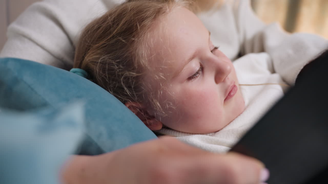 Child resting on soft pillow while looking up toward mother during quiet reading moment, creating intimate family scene filled with comfort, care, emotional warmth, and close nurturing atmosphere in cozy home
