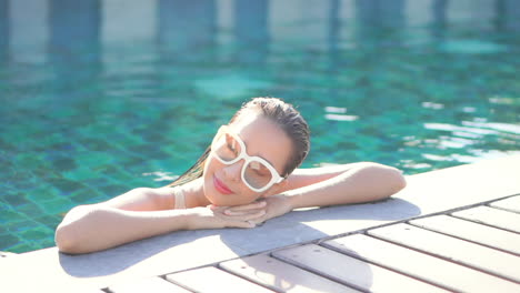 face closeup of a beautiful asian woman in white sunglasses relaxing in swimming pool daytime putting down her head on locked hands