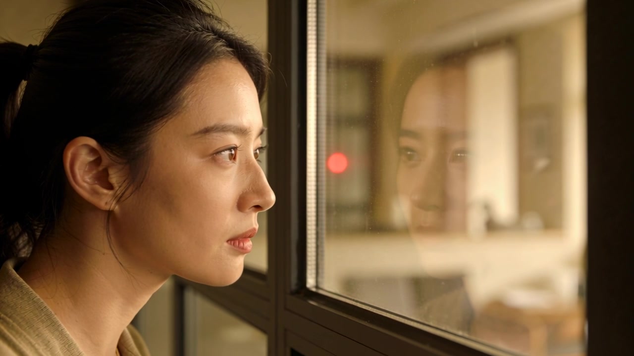 방 안에서 기대어 서 있는 아시아 여성이 유리 반사면에 얼굴을 맞추고, 붉은 빛을 알아차린 후 눈을 감는 모습
