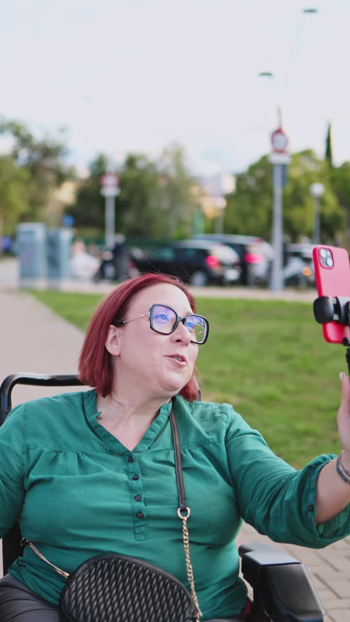 Donna felice in sedia a rotelle che fa vlogging con lo smartphone al parco.