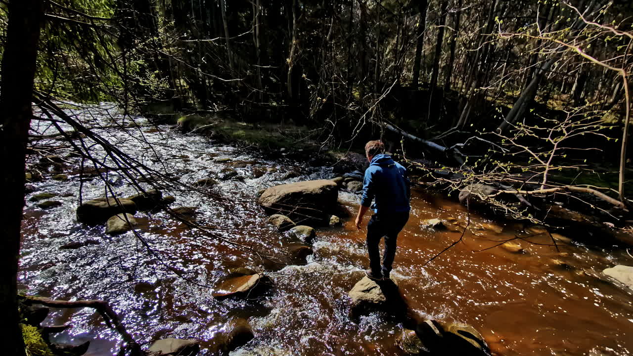 A Man Leaps Across a Rocky Stream in a Sunlit Forest, Capturing a Moment of Adventure in Nature in Riga, Latvia - High Angle Shot