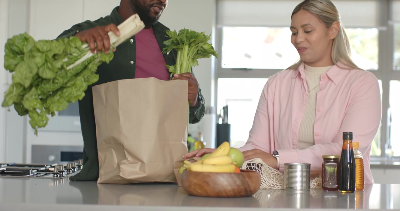 Pareja afroamericana en la cocina; el hombre coloca una bolsa y clasifica verduras mientras ella embolsa manzanas para almacenar.