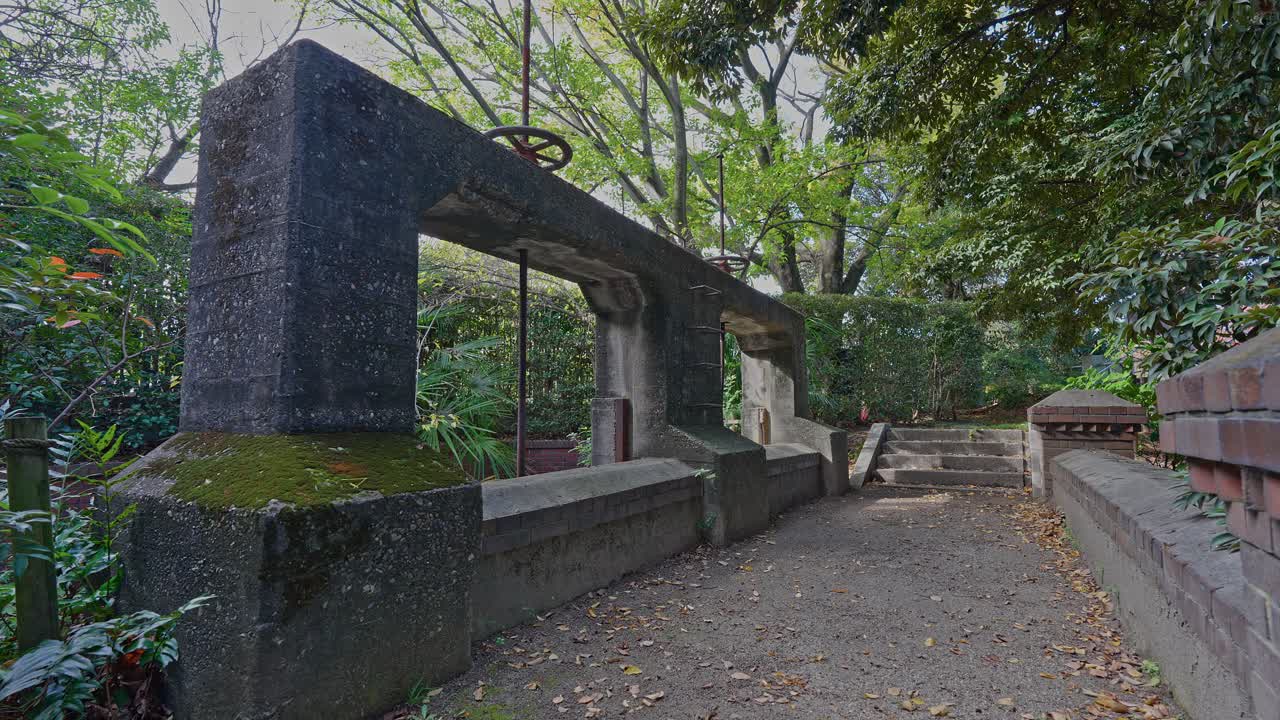 Historic concrete irrigation weir gate (Jinzaemon Weir) with a visible valve wheel, surrounded by overgrown park greenery