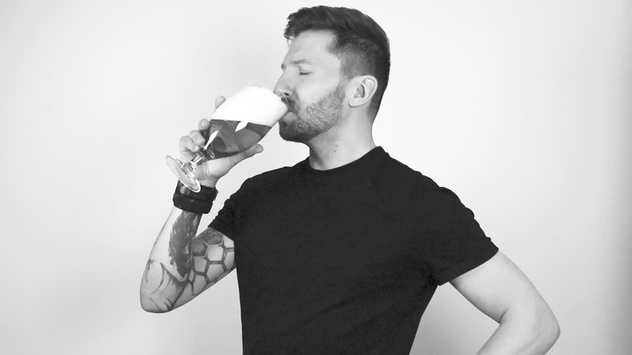 imágenes en escala de grises de un hombre tatuado levantando un vaso de cerveza con espuma en la parte superior y bebiendo de él. después de beber asintió con la cabeza con aprobación indicando que le gustaba. el hombre tiene el cabello corto y una barba.