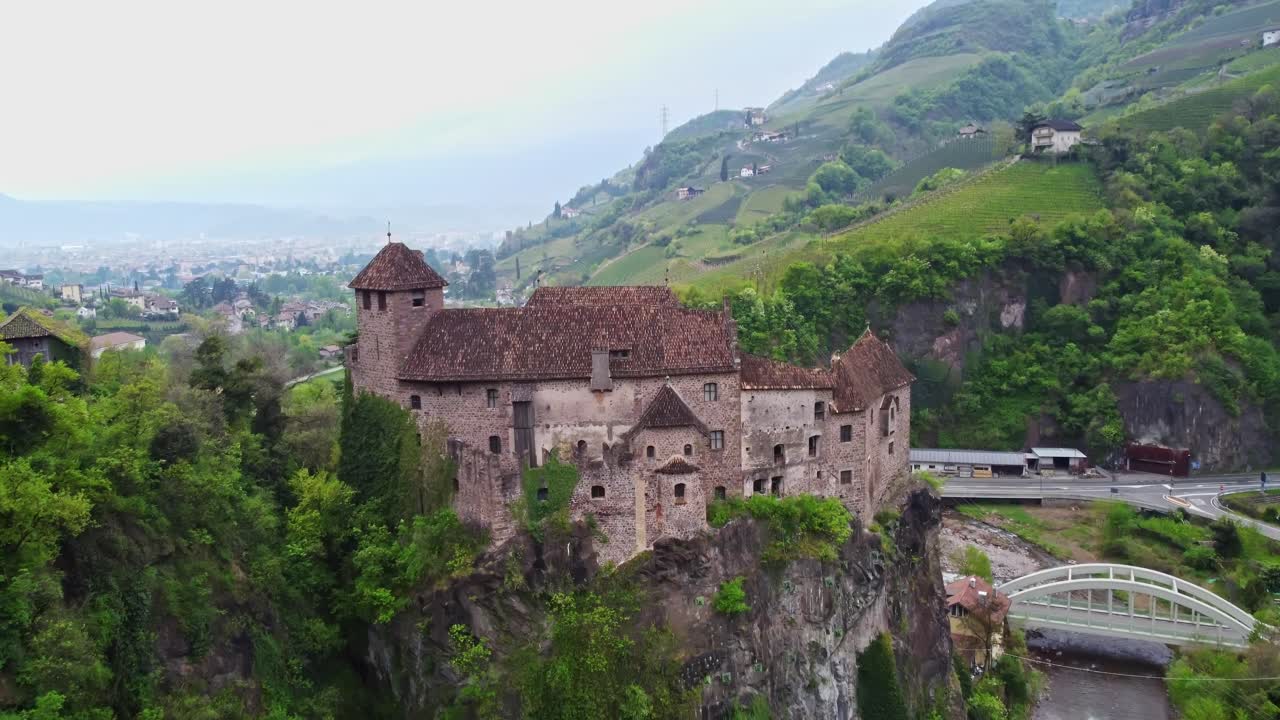Drone ascends alongside Castel Roncolo revealing its medieval structure over the Talvera river gorge and sloped vineyards of Renon near Bolzano in Trentino-Alto Adige, slow motion