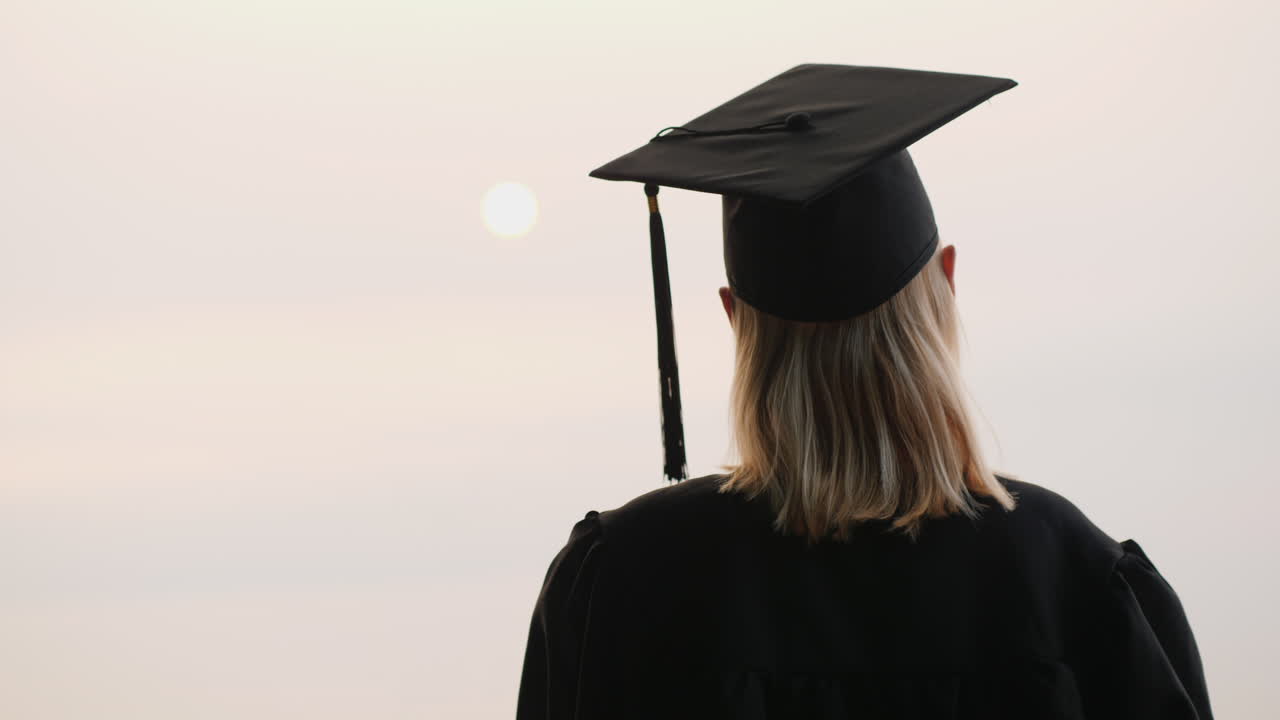 un graduado con manto y gorra mira el amanecer sobre la vista trasera del mar