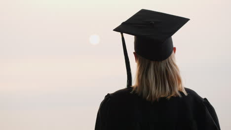 a graduate in a mantle and cap looks at the sunrise over the sea rear view