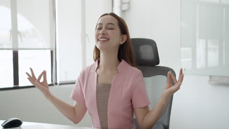A smiling woman in a meditative formation relaxes her mind while sitting at her office.