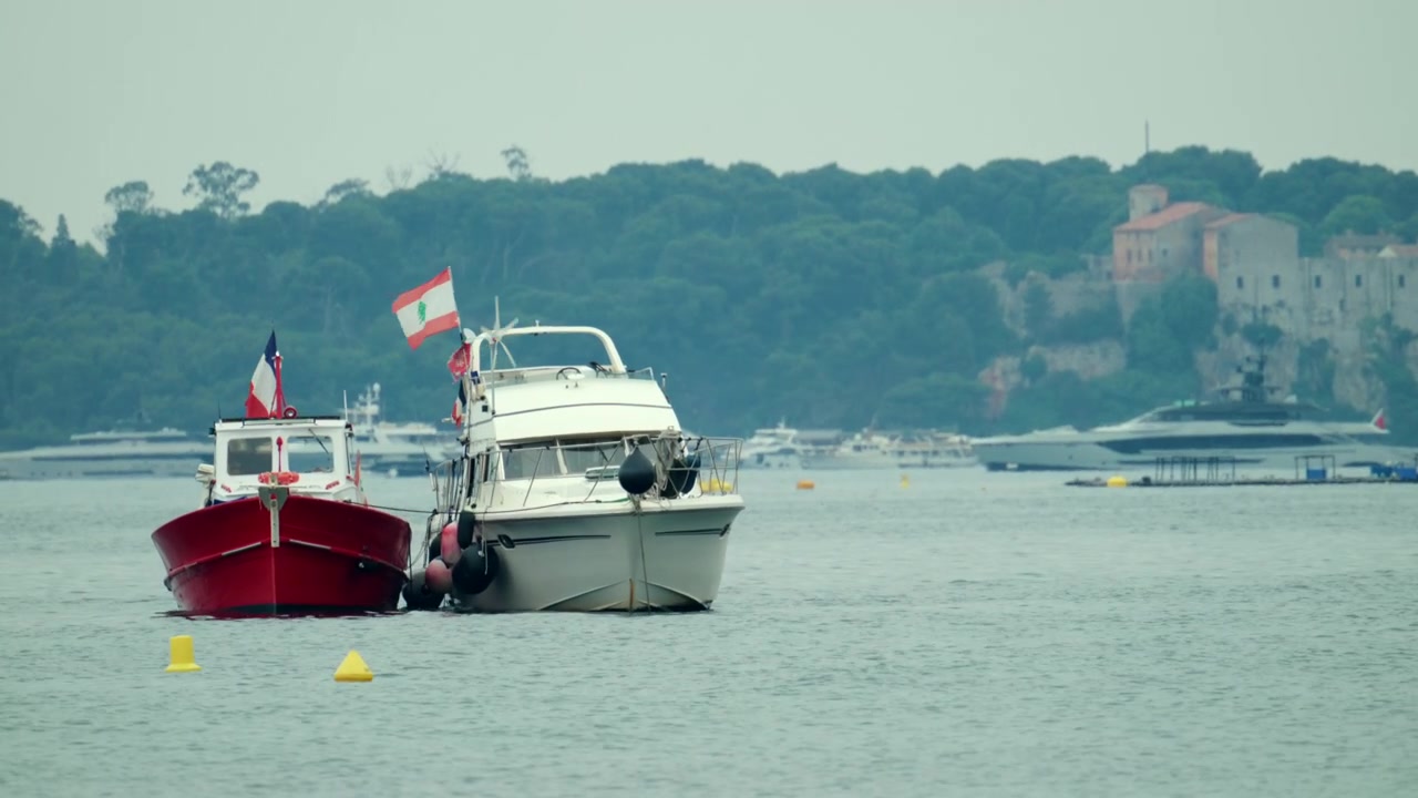 Boote ankern in einer ruhigen Bucht, mit den Nationalflaggen des Libanon und Frankreichs, einer Küstenlinie und einem historischen Gebäude im Hintergrund.