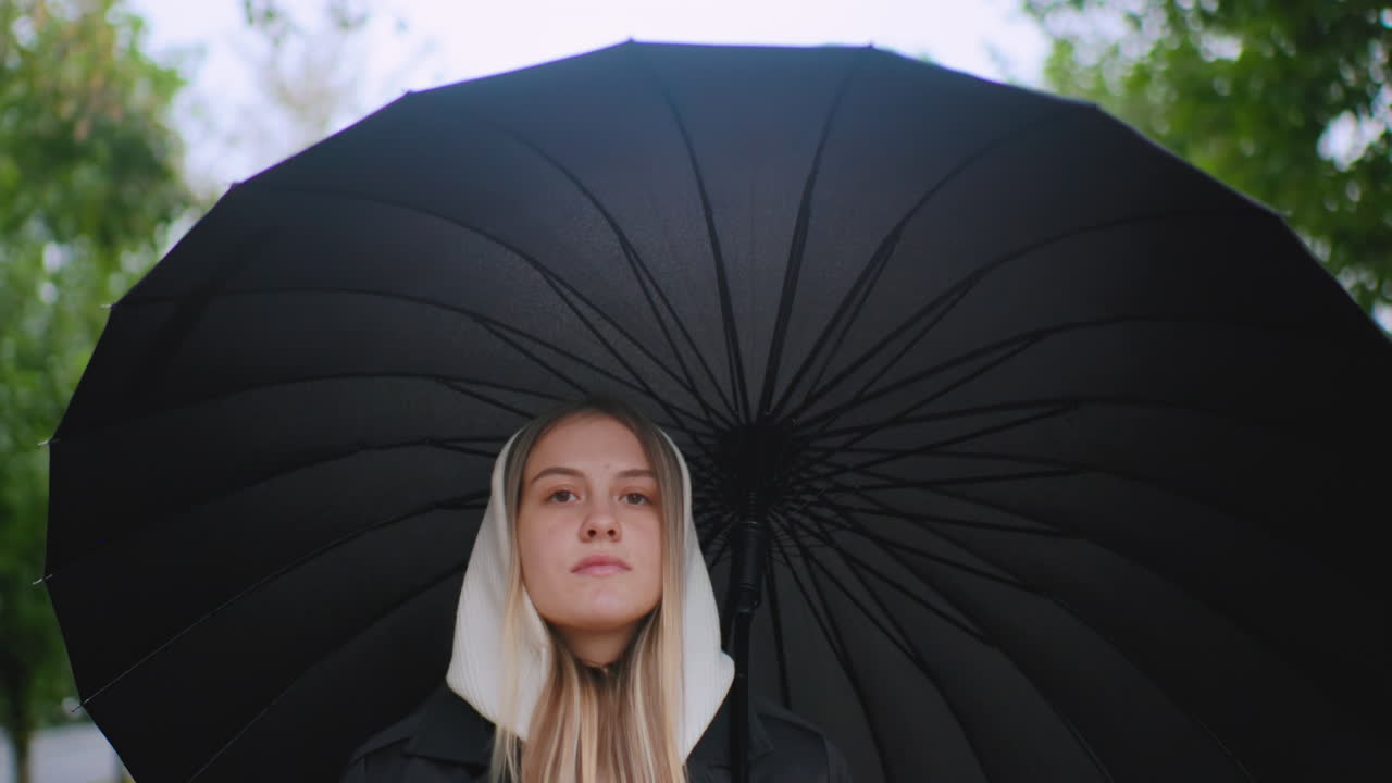 Young woman in white hood walks under large black umbrella outdoors, calm expression on face, moody weather atmosphere, natural light highlighting hair , urban park background with green blurred trees