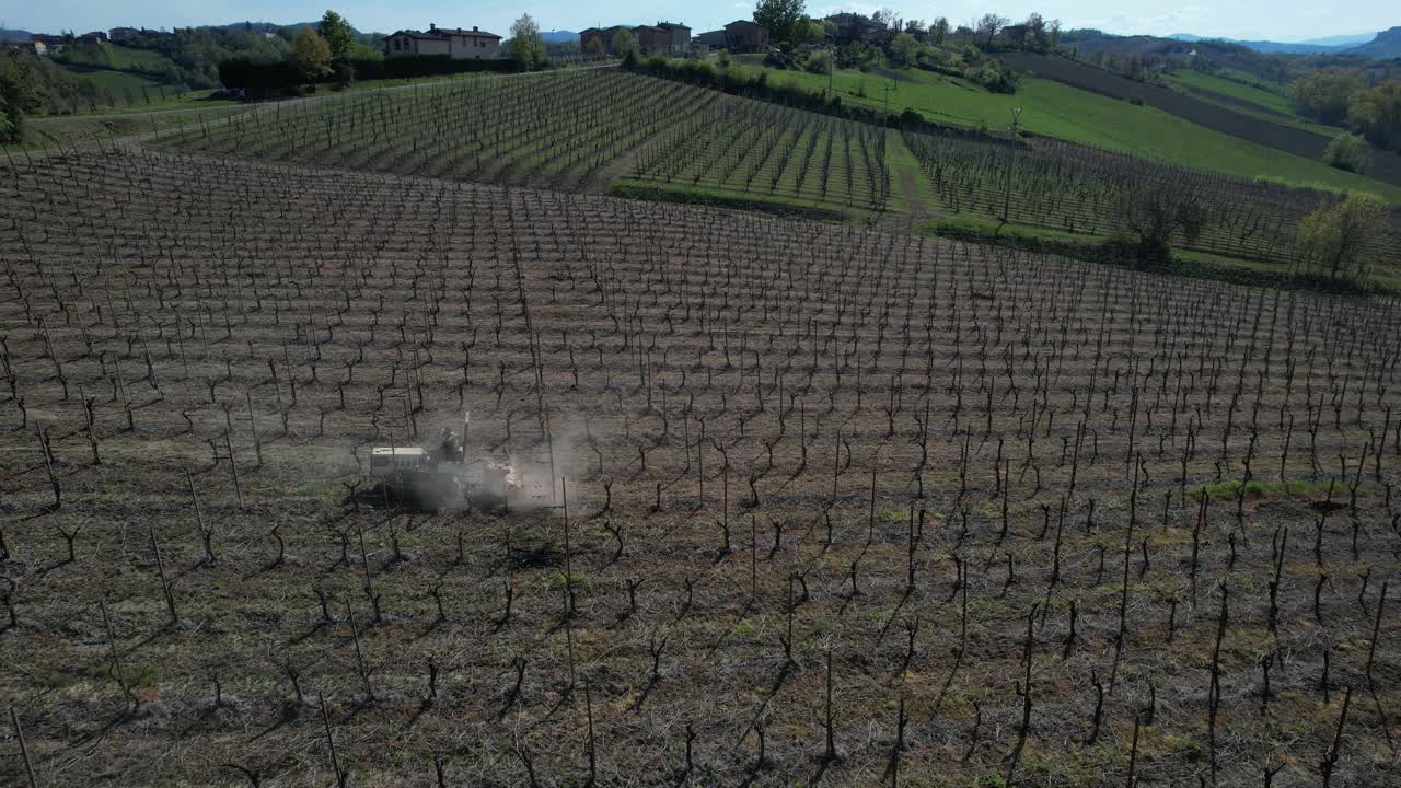 Trattore agricolo esegue diserbo meccanico lungo filari di vigneto allineati in un campo coltivato all'inizio della stagione vegetativa, ripresa da drone