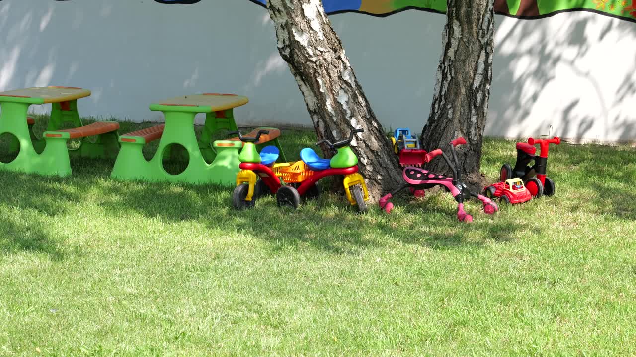 Jouets à chevaucher, camions et tables de pique-nique en plastique sous un arbre devant une école primaire
