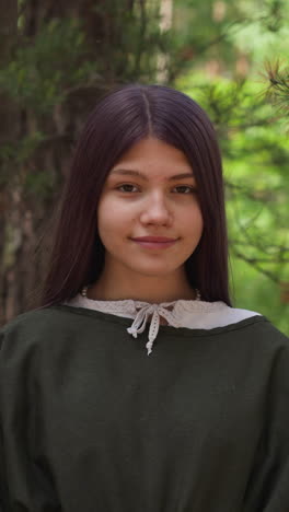 cheerful teenage girl with purple hair in clothes of middle age period stands in green forest closeup. participant of history reconstruction camp