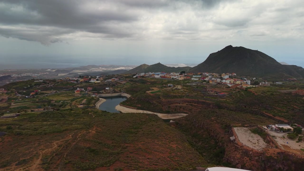 Veduta aerea di un terreno vulcanico aspro, un villaggio e un bacino idrico sotto cieli nuvolosi.