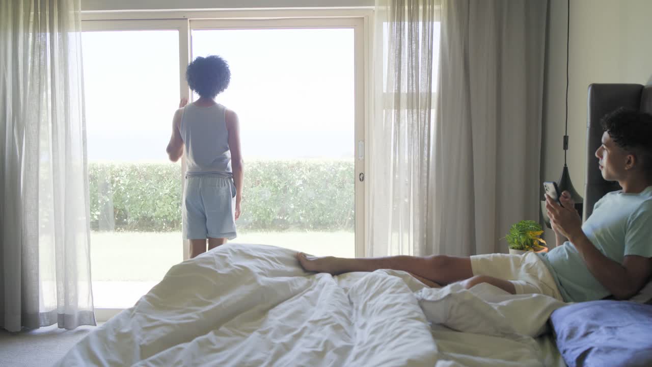 Partner maschi afroamericani, uno che tira le tende mostrando il cortile mentre l'altro guarda il telefono sul letto
