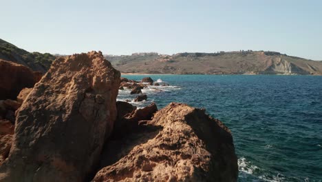 Flying-slow-with-a-drone-above-seashore-with-beautiful-turquoise-waters-of-the-Mediterranean-Sea-splashing-on-the-red-coloured-rocks