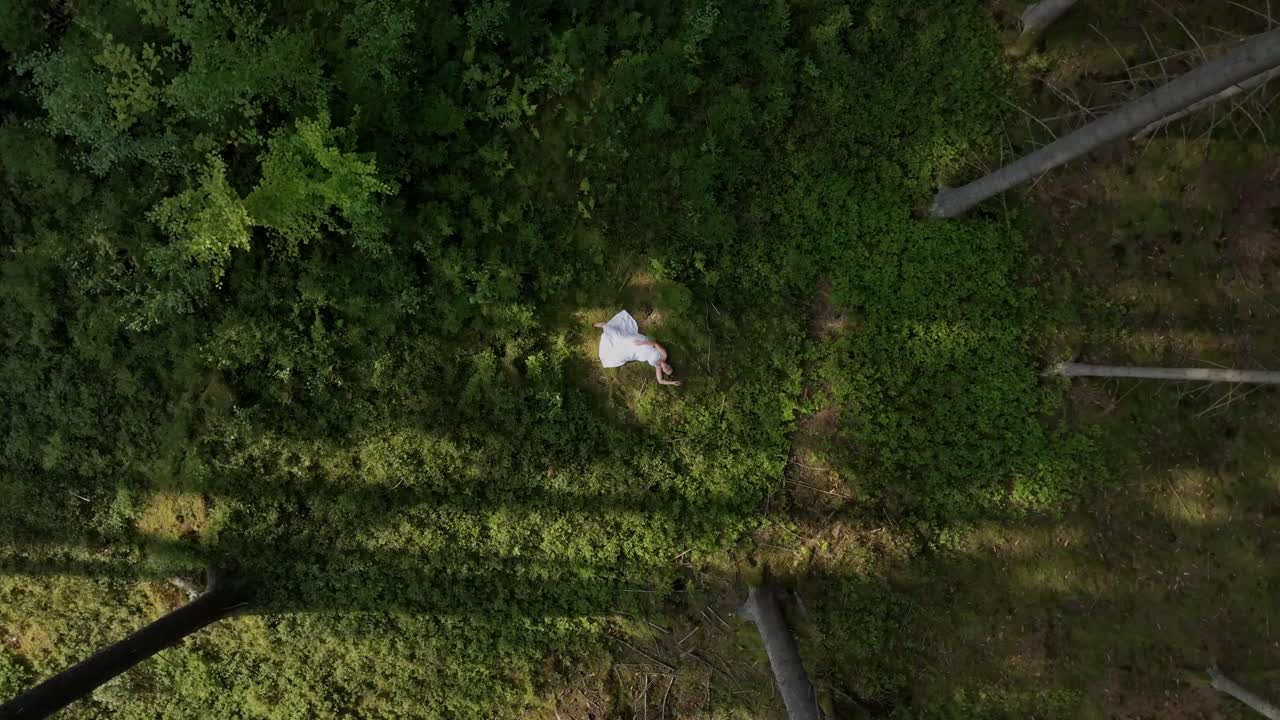 Woman in white dress lying in a forest, viewed from above, embracing nature in peaceful harmony and relaxation.