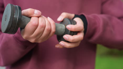 close view shows sports woman in wine hoodie gently tightening end cap on dumbbell, palms securing handle during outdoor strength training near courtyard, highlighting focus and muscle care
