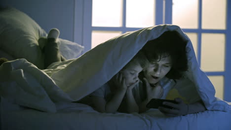 cute little girl and young mother lying on bed under blanket and reading fairy tale with smartphone together