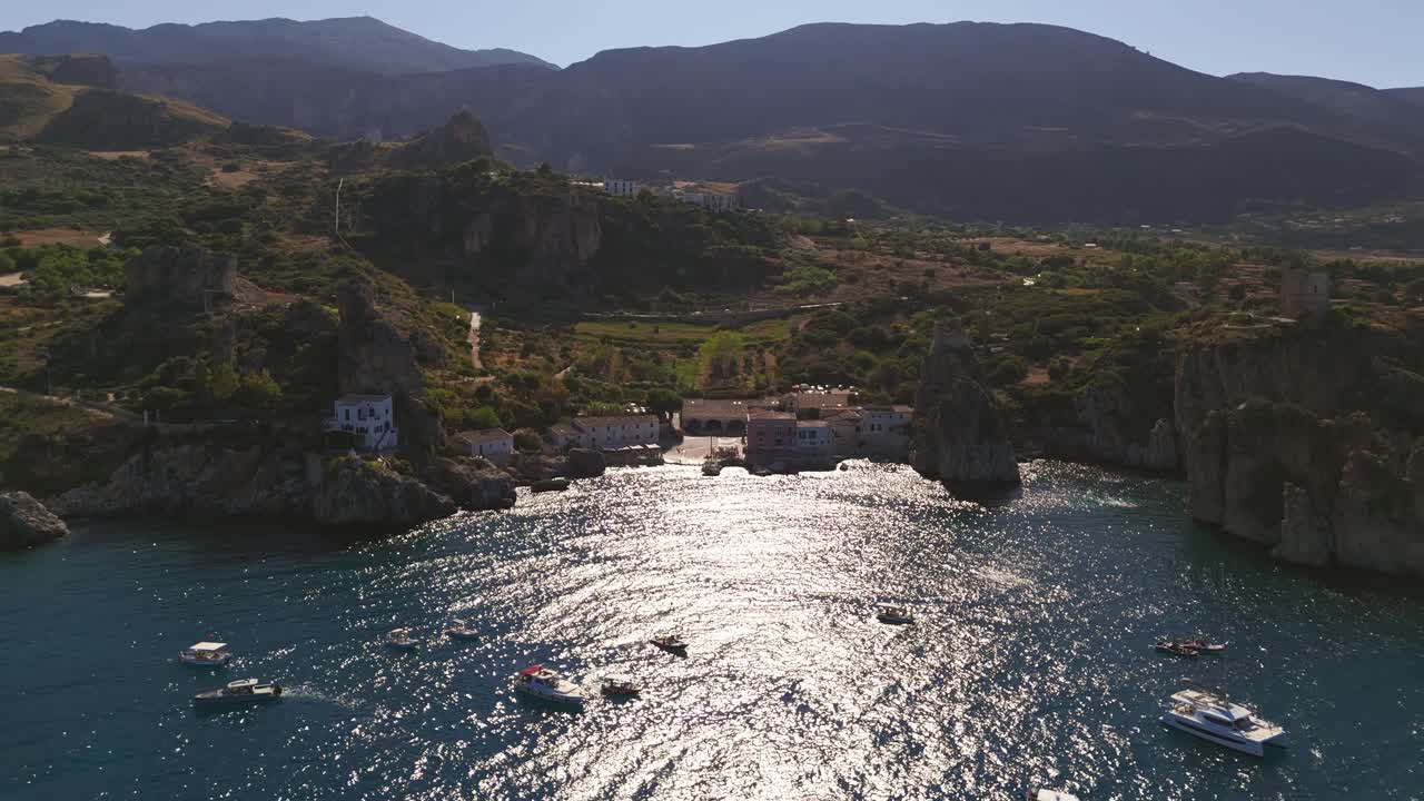 Aerial drone shot pulling back above the crystal-clear waters of Tonnara di Scopello, Sicily, Italy, unveiling numerous anchored boats surrounded by the stunning coastal cliffs and turquoise sea