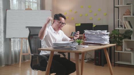 happy asian man celebrating winning game on smartphone after working with documents at the office