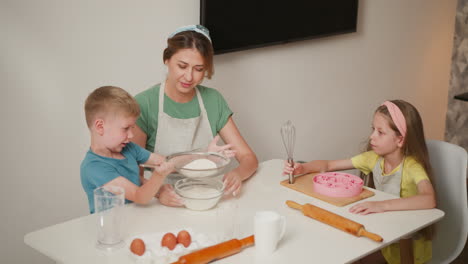 overhead view of young boy sieving flour into bowl held by woman in apron while little girl sits nearby holding whisk, surrounded by baking tools including eggs, rollers, and pink molds on table
