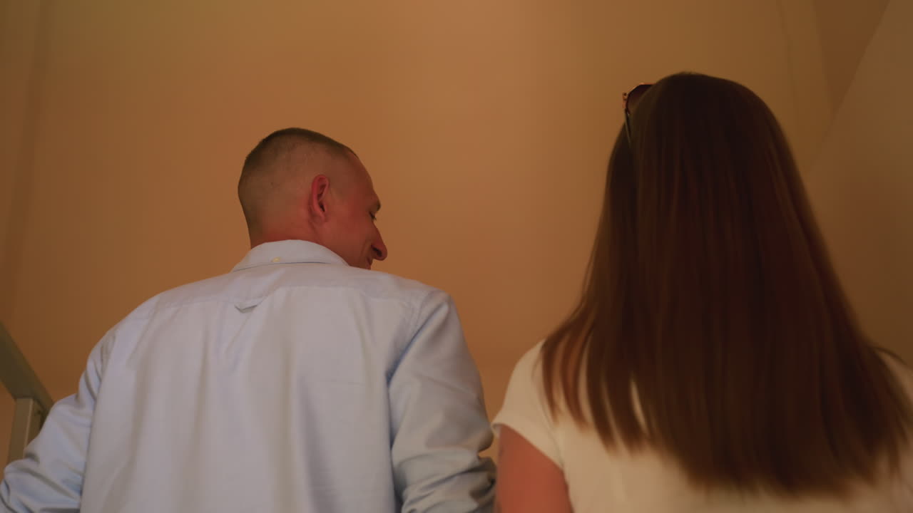 Pareja subiendo las escaleras dentro de un apartamento, vista trasera de un hombre con camisa clara y una mujer con top blanco con tatuaje visible en el brazo, pared beige neutra e interior vacío, inspección y planificación silenciosas