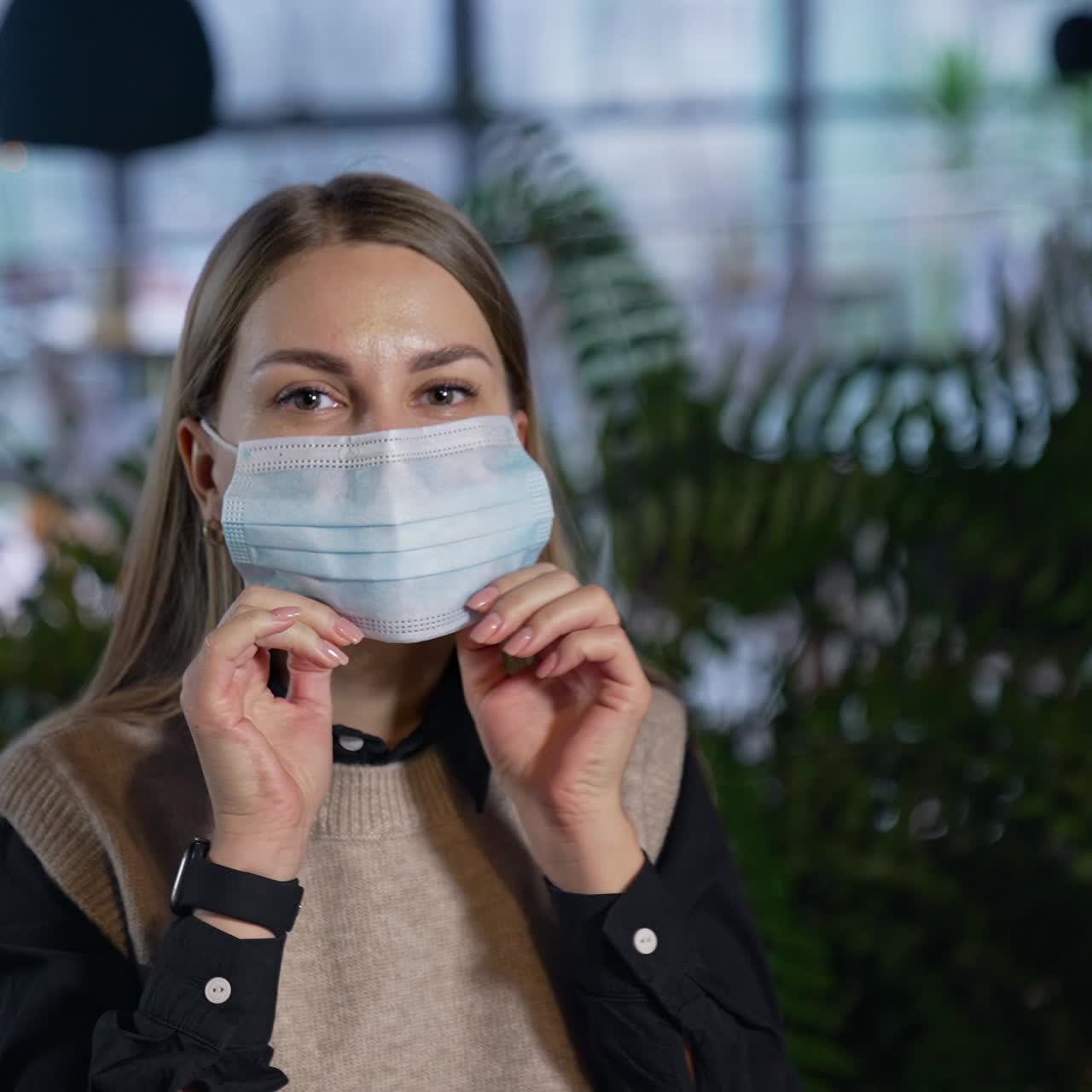 Attractive female office worker puts on mask. Lady in mask looks straight into the camera. Woman in mask against the blurred backdrop