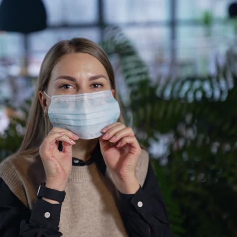 attractive female office worker puts on mask. lady in mask looks straight into the camera. woman in mask against the blurred backdrop